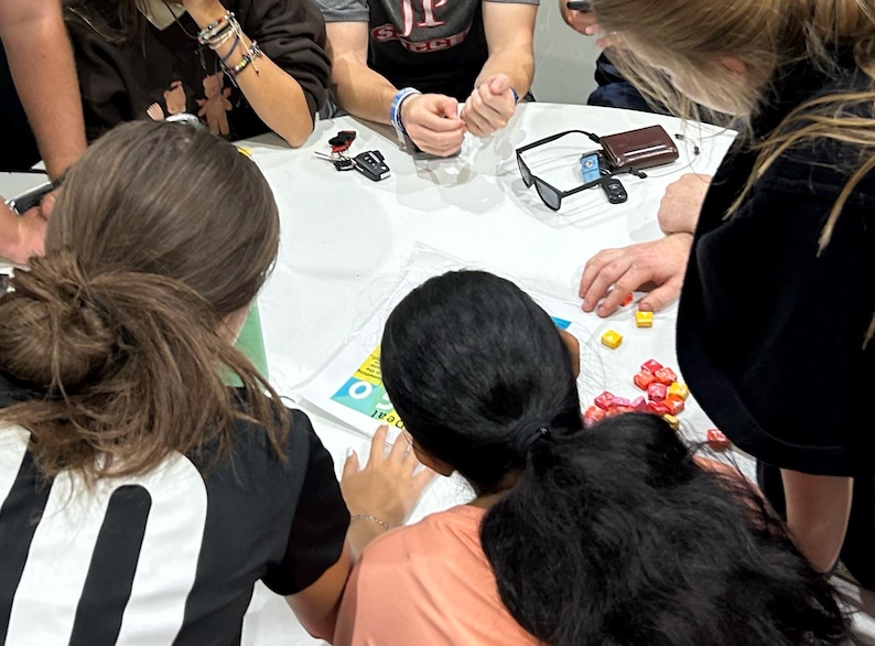 May include: A group of people are gathered around a table, working on a project. They are using colourful tiles to create a design. The table is covered in a white tablecloth.