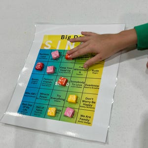May include: A colourful game board with the title "Big D Sing" and a grid of song titles. The game board has a yellow, green and blue colour scheme. There are small, colourful candies on the board.