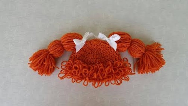 May include: A bright orange crocheted hat featuring two thick yarn pigtails on either side, each tied with a small white ribbon bow. The hat has a textured, looped fringe along the bottom edge, mimicking a hairstyle with bangs and side braids.
