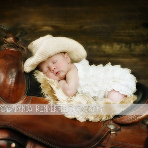 May include: A baby girl wearing a white lace dress and a cream-colored cowboy hat sleeps on a brown leather saddle.