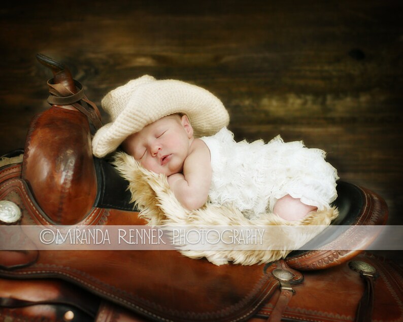 May include: A baby girl wearing a white lace dress and a cream-colored cowboy hat sleeps on a brown leather saddle.
