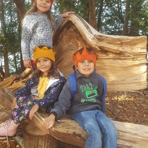 Puede incluir: Tres niños con gorros de ganchillo de colores posan en un entorno boscoso. Los niños están sentados en un tronco de árbol caído. La niña de la izquierda lleva un gorro rojo, la niña del medio lleva un gorro amarillo y el niño de la derecha lleva un gorro naranja.