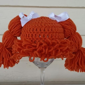 May include: A bright orange crocheted hat featuring thick yarn pigtails on each side, each tied with a white ribbon bow. The hat includes a textured fringe of orange yarn across the front, mimicking bangs, and is constructed with a dense, looped stitch pattern.