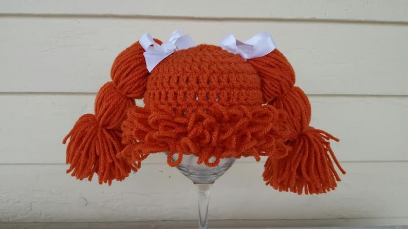 May include: A bright orange crocheted hat featuring thick yarn pigtails on each side, each tied with a white ribbon bow. The hat includes a textured fringe of orange yarn across the front, mimicking bangs, and is constructed with a dense, looped stitch pattern.
