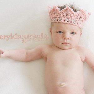 May include: A baby wearing a pink crocheted crown, lying on a white surface. The baby has a serious expression and is looking directly at the camera.