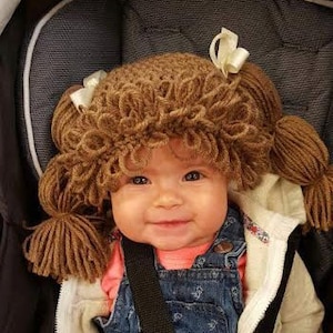 May include: A brown yarn hat designed to resemble hair with looped bangs and two side pigtails tied with small white ribbons. The hat features a textured, curly appearance made from thick brown yarn, creating a playful look that covers the head.
