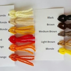 May include: A color chart featuring twelve yarn samples attached to two white cards. The left card lists Light Blonde, Dirty Blonde, Golden Blonde, Red, Orange, and Carrot Orange. The right card lists Black, Brown, Medium Brown, Light Brown, and Blonde.