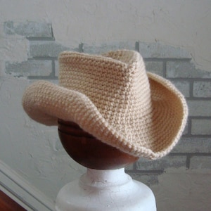 May include: A crocheted cowboy hat in a cream color. The hat is sitting on a white pedestal.