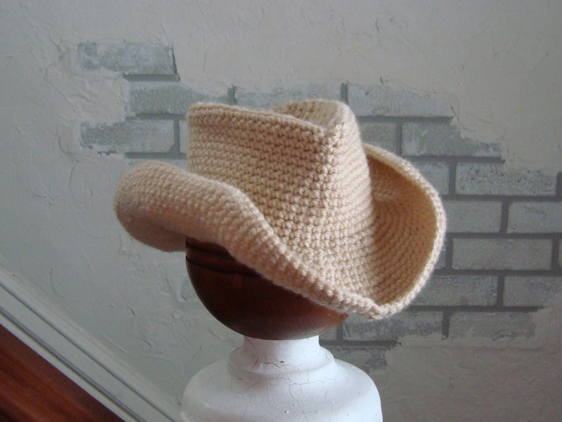 May include: A crocheted cowboy hat in a cream color. The hat is sitting on a white pedestal.