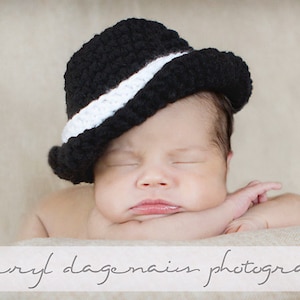 May include: A newborn baby wearing a black and white crocheted hat with a brim. The baby is sleeping with their arms crossed under their chin.