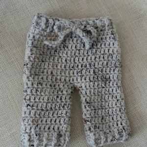 Puede incluir: Un pantalón de bebé de ganchillo de color gris claro con motas marrones y negras. El pantalón tiene una cintura con cordón.
