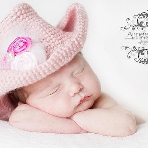 May include: A sleeping baby girl wearing a pink crocheted cowboy hat with a pink and white flower embellishment.