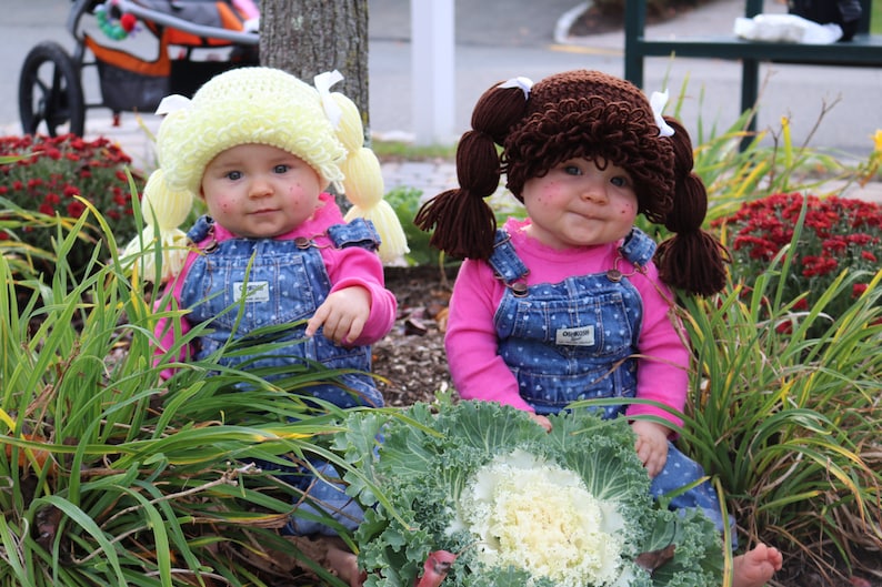 May include: Two crochet yarn wigs, one in pale yellow and one in dark brown, styled with pigtails and white bows. The wigs are designed to resemble doll hair, featuring textured loops and yarn strands, worn over the heads of two small children.