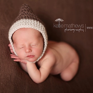 May include: A newborn baby wearing a brown and white knitted hat with a pointed tip, sleeping on a brown textured surface.