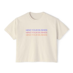 May include: A cream-coloured t-shirt featuring the phrase "MIND YOUR BUSINESS" in a layered gradient of blue, purple, and pink. The text is repeated three times. The shirt has a crew neck and short sleeves. A casual, everyday garment.