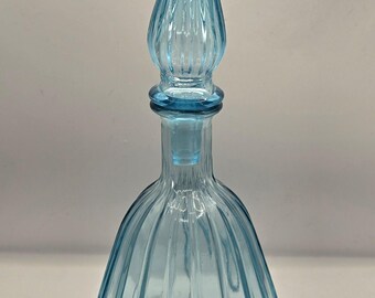 May include: A light blue glass bottle with a tall, pointed stopper. The bottle has a ribbed design and a rounded base. The stopper is also ribbed and tapers to a point. The bottle is empty and sits on a white surface.