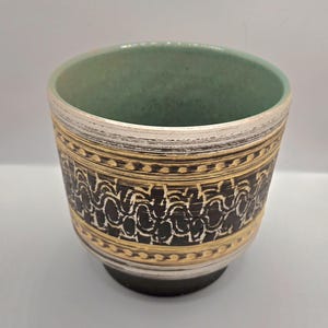 Puede incluir: Un macetero de cerámica con un interior esmaltado verde y un diseño geométrico negro y dorado. El macetero tiene una base blanca con un borde negro.
