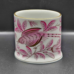 May include: White ceramic mug with a pink bird and floral design. The bird is depicted in flight with pink and brown details. The leaves and flowers are in shades of pink. The rim has a gold trim.