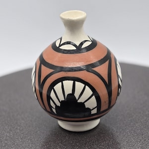 May include: A small, round, ceramic vase with a geometric design. The vase is painted in black, white, and pink.