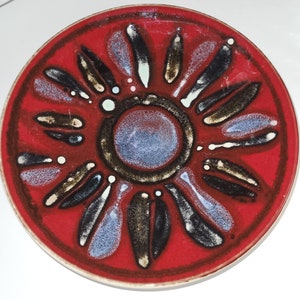 Puede incluir: Un plato de cerámica rojo con un diseño de flor estilizado en blanco y negro. La flor tiene un círculo central con un centro azul y blanco.
