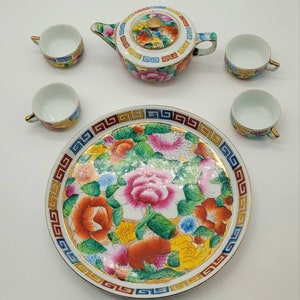 May include: A miniature tea set with a floral design. The set includes a teapot, four teacups, and a plate. The teapot has a lid with a floral design and a gold trim. The teacups and plate also have a floral design and gold trim.