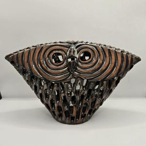 Puede incluir: Un cuenco decorativo con forma de búho y un diseño calado detallado. El cuenco presenta una combinación de colores marrón y negro, con círculos concéntricos que forman los ojos del búho. El calado permite el paso de la luz.