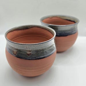 May include: Two terracotta mugs with a dark blue and grey glaze. The mugs are round and have a slightly flared lip.