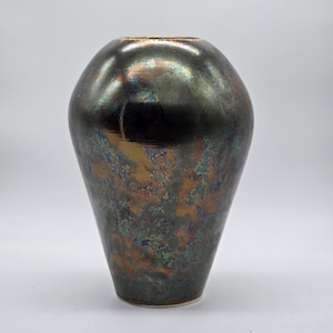 May include: A dark, iridescent ceramic vase with a rounded body and a slightly flared base. The vase has a metallic finish with shades of green, bronze, and blue. The surface appears textured, with a glossy sheen. The vase is a decorative home decor item.