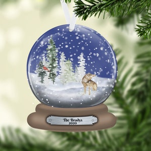 May include: A blue glass snow globe ornament with a winter scene inside. The scene includes a snowy forest with pine trees, a red cardinal, and a wolf. The base of the globe is brown with a silver plaque that reads "The Bradys 2020".