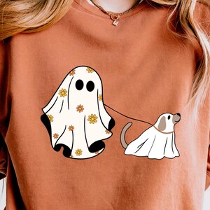 May include: A rust-colored sweatshirt with a white ghost and a white dog wearing a ghost costume. The ghost has a floral pattern and is holding a leash attached to the dog.