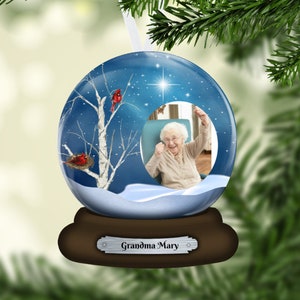 May include: A blue glass snow globe ornament with a winter scene inside. The scene includes a birch tree, two red cardinals, and a photo of a woman smiling. The base of the snow globe is brown wood with a silver plaque that reads "Grandma Mary".