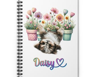 May include: A white spiral notebook with a colorful illustration of a puppy upside down in a flower pot. The puppy is brown and white with a black nose. The flower pot is decorated with pink and green stripes. The flowers are pink, white, and yellow. The text "Daisy" is written in rainbow colors below the puppy.
