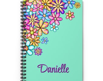 May include: A turquoise spiral notebook with a colorful floral design in the top corner. The name Danielle is written in purple cursive at the bottom of the notebook.