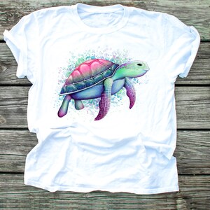 May include: White t-shirt featuring a watercolour illustration of a sea turtle in shades of pink, blue and green.