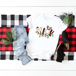 May include: White t-shirt with a Christmas-themed design featuring a pair of scissors, a hairbrush, a curling iron, and a comb. The design is surrounded by a garland of lights.