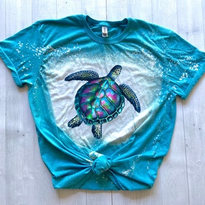 May include: A turquoise blue t-shirt with a bleach-splattered design featuring a colourful sea turtle graphic.