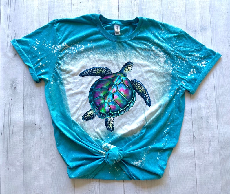 May include: A turquoise blue t-shirt with a bleach-splattered design featuring a colourful sea turtle graphic.