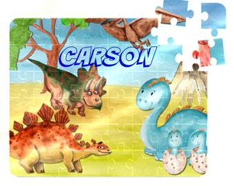 May include: A colorful dinosaur-themed jigsaw puzzle with the name "CARSON" printed in blue letters. The puzzle features a green and brown triceratops, a brown stegosaurus, and a blue brachiosaurus with two baby dinosaurs.