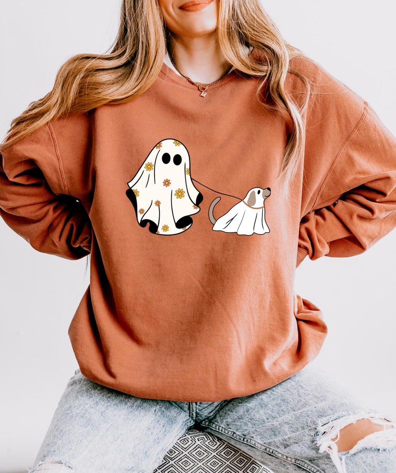 May include: A rust-colored sweatshirt with a white ghost and a white dog wearing a ghost costume. The ghost has a floral pattern and is holding a leash attached to the dog.