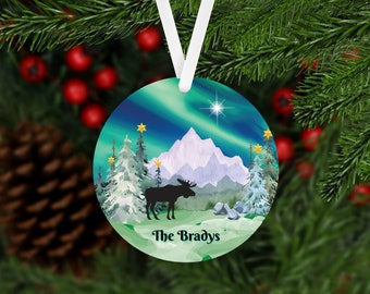 May include: Round Christmas ornament with a green, blue, and white winter scene. The ornament features a silhouette of a moose standing in front of a snowy mountain range with a green forest in the foreground. The aurora borealis is visible in the sky above the mountains. The text "The Bradys" is printed in black at the bottom of the ornament.