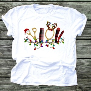 May include: White t-shirt with a Christmas-themed design featuring a pair of gold scissors, a comb, a round mirror, a red and black plaid hair cutting cape, and a Santa hat. The design is surrounded by a string of colorful Christmas lights.