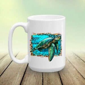 May include: White ceramic mug featuring a detailed illustration of a sea turtle. The turtle is depicted in shades of green and blue, set against a turquoise background with a leopard print border. The mug has a large handle.