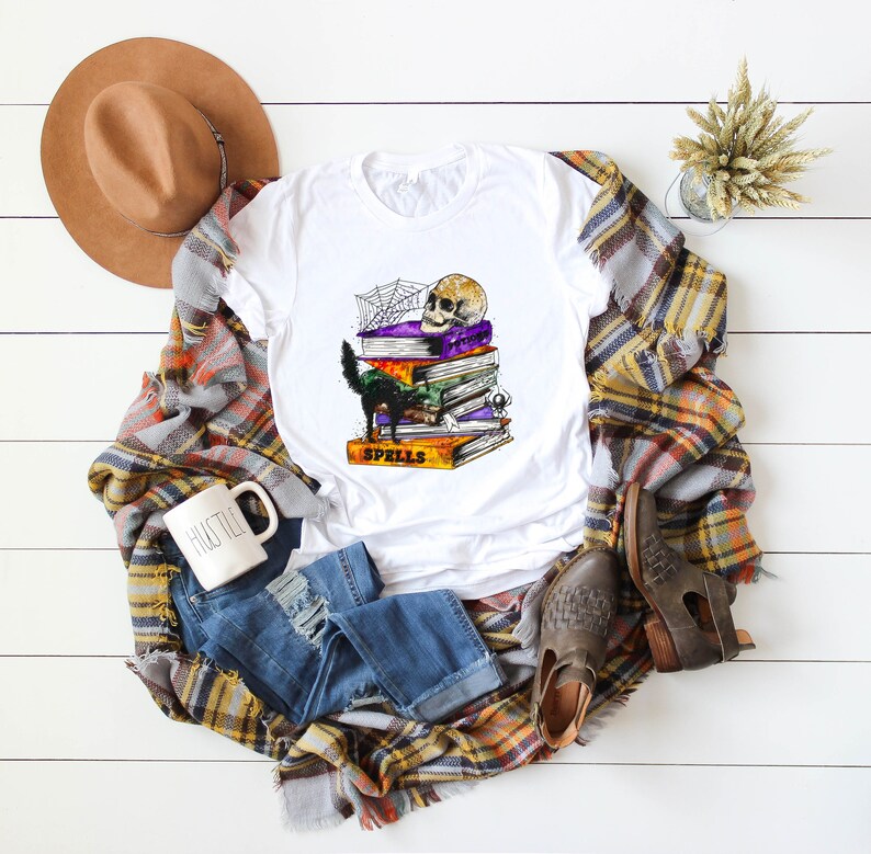 Peut inclure: T-shirt blanc avec un chat noir, un cr&acirc;ne et une pile de livres avec le mot "Spells" sur le livre du bas. Les livres sont d&eacute;cor&eacute;s d'une toile d'araign&eacute;e et d'autres d&eacute;tails sur le th&egrave;me d'Halloween.