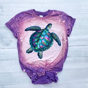 May include: A purple and pink tie-dye t-shirt with a bleach-splattered design featuring a colourful sea turtle graphic.