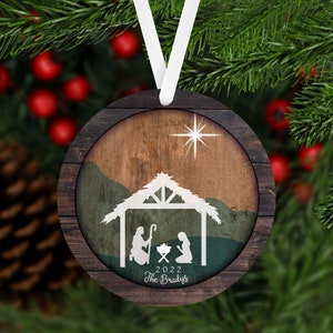 May include: A round wooden Christmas ornament with a rustic woodgrain design. The ornament features a white silhouette of the nativity scene with a star above the stable. The text "2022 The Bradys" is printed below the scene.