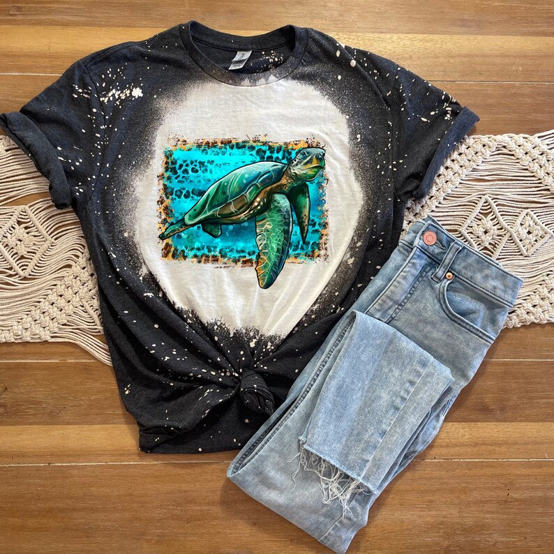 May include: A dark gray t-shirt with a bleached white circle design, featuring a turquoise and green sea turtle graphic. Light blue jeans are folded next to the shirt. The shirt is knotted at the waist.