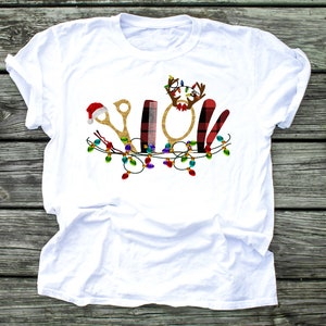 May include: White t-shirt with a Christmas-themed design featuring a pair of gold scissors, a comb, a round mirror, a red and black plaid hair cutting tool, and a Santa hat. The design is surrounded by a string of colorful Christmas lights.