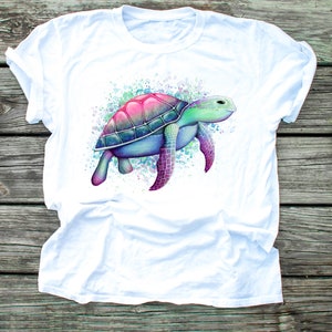 May include: White t-shirt featuring a watercolour illustration of a sea turtle in shades of pink, blue and green.