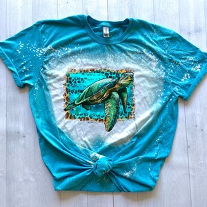 May include: A turquoise t-shirt with a bleached design and a sea turtle graphic. The turtle is green and brown, set against a turquoise and leopard print background. The shirt is knotted at the bottom.