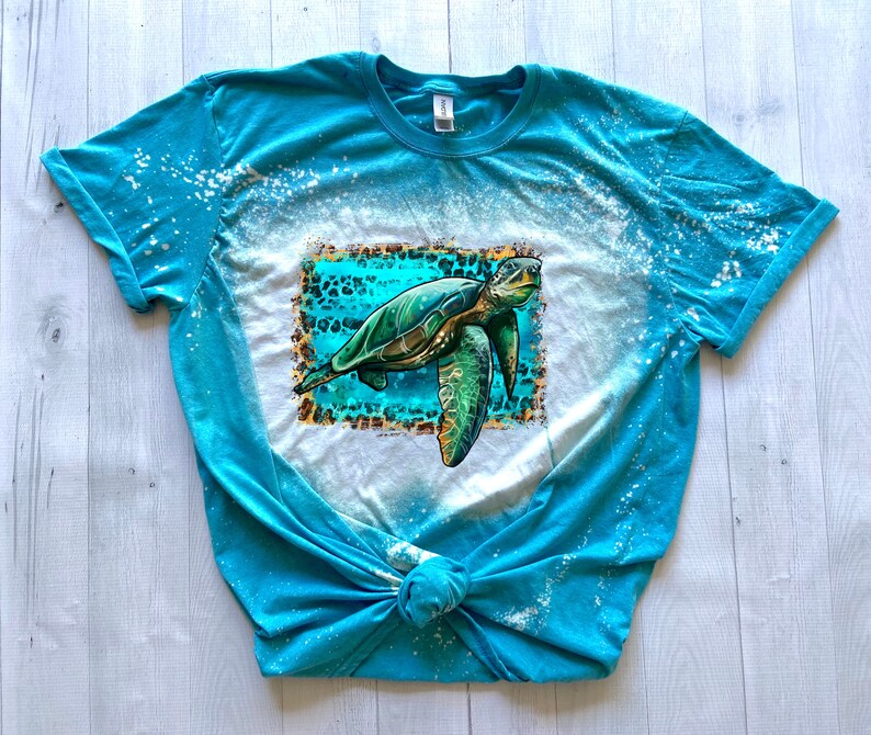 May include: A turquoise t-shirt with a bleached design and a sea turtle graphic. The turtle is green and brown, set against a turquoise and leopard print background. The shirt is knotted at the bottom.